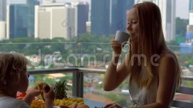 年轻的女家庭教师和一个男孩在阳台上吃早餐，俯瞰市中心的摩天大楼